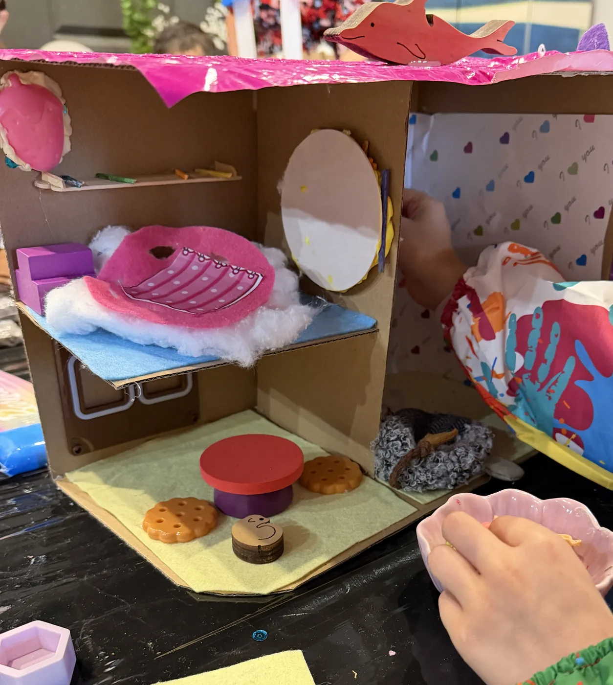 A cardboard doll's house furnished inside with a bed, a table and paper decorations