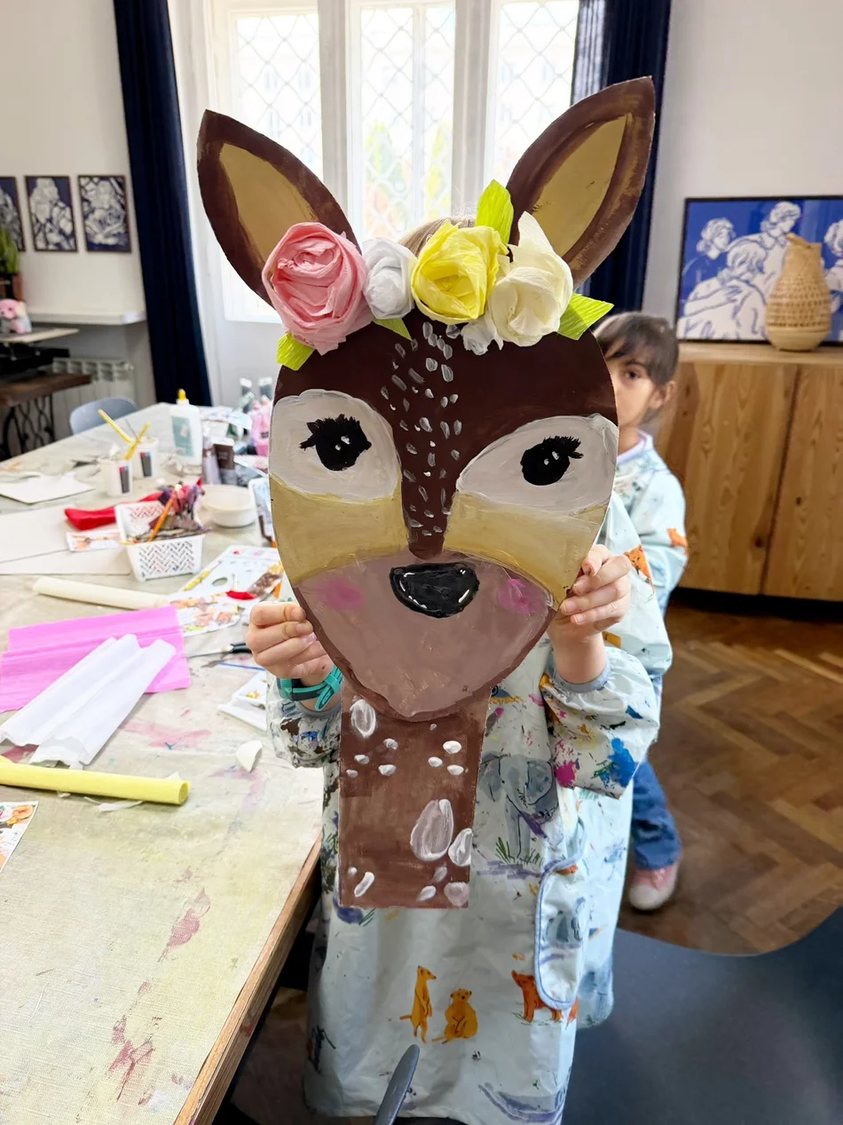 A child holding up a painted deer mask with a paper flower crown