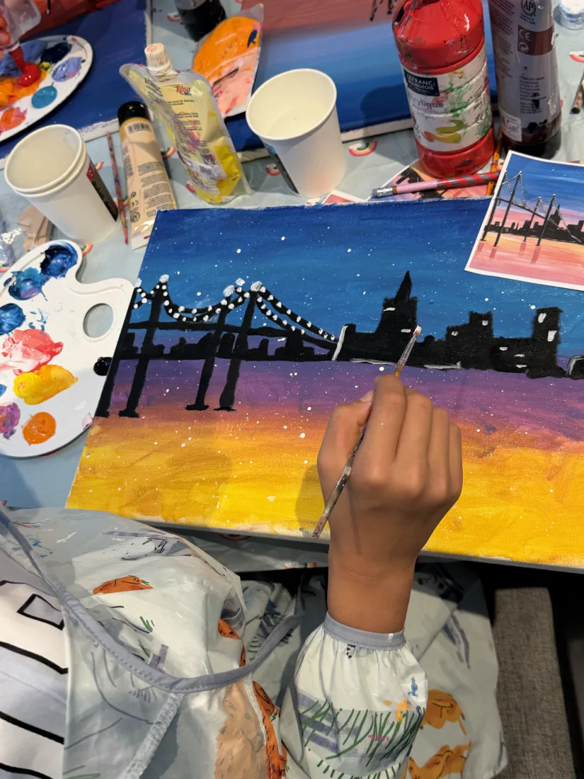 A child painting a night-time city skyline with a bridge and lights on canvas